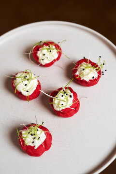 Close-up of elegant red choux pastry appetizers filled with white mousse, garnished with sprouts and sesame on a white plate. Gourmet catering, buffet, banquet, or wedding reception scene