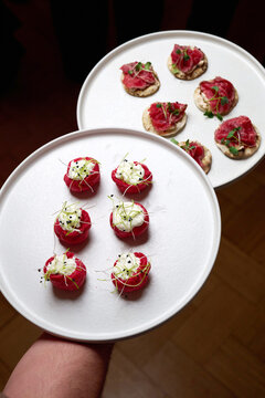 Close-up of a waiter holding two white plates with gourmet appetizers. Featuring red choux pastry with white mousse and roast beef crostini. Elegant catering for a banquet or wedding reception