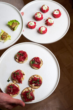 Close-up of a waiter holding three white plates with gourmet appetizers. Featuring red choux pastry with white mousse and roast beef crostini. Elegant catering for a banquet or wedding reception