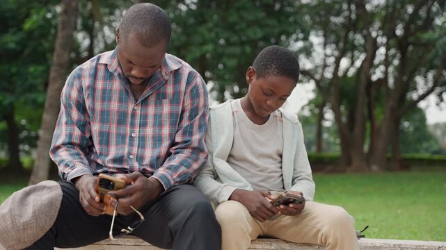 Black father and son sitting bench, both focused on phones, gentle companionship in leafy park, casual clothes, father mentoring son through simple technology, calm afternoon mood, trees and green