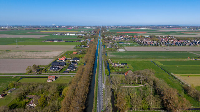 Aerial view of a long straight road and canal running through flat agricultural fields and residential areas under a clear blue sky in the Netherlands.