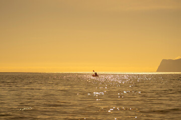 Fototapeta premium Kayaking, sunset, ocean. Person paddles red kayak on sparkling golden water with distant mountains.