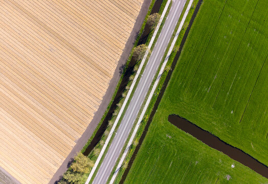 Aerial view of a straight asphalt road dividing a ploughed agricultural field and a lush green meadow with narrow drainage canals in Heiloo, North Holland, Netherlands.