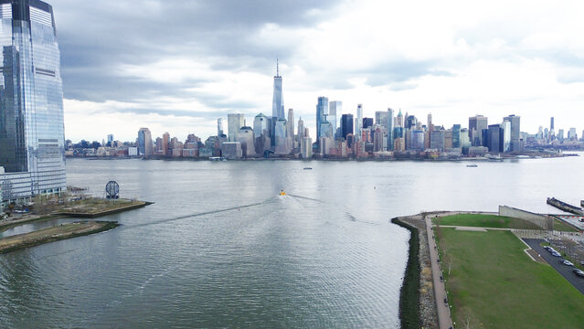 Riverfront skyline sweeps capture Hudson River edge near Liberty State Park facing Lower Manhattan tower cluster, One World Trade Center rising. Wake trails, reflective facades, heavy cloud, NJ
