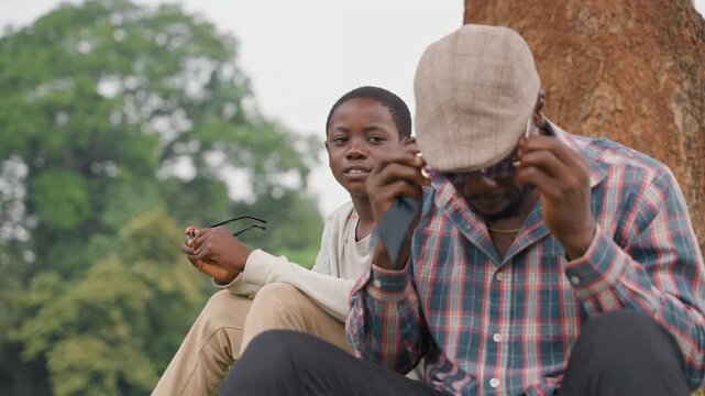 Black father and son closeup sitting by tree, father adjusts glasses and inspects device while son watches attentively, quiet mentorship vibe, patterned shirt and cap, green nature background
