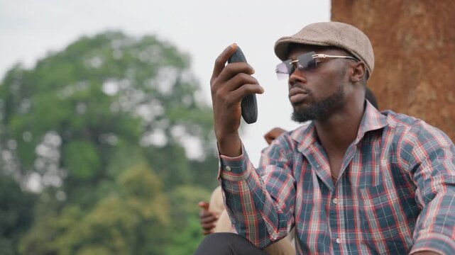 Black father and son outdoors by tree, father holds handheld radio up and gestures while son listens closely, storytelling and teaching vibe, plaid shirt and cap, green park background and focused