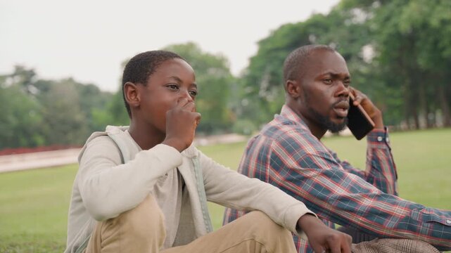 Black father and son with father on phone and son speaking up, park grass and trees backdrop, father balancing work call while son asserts opinion, moment of negotiation, candid lifestyle portrait