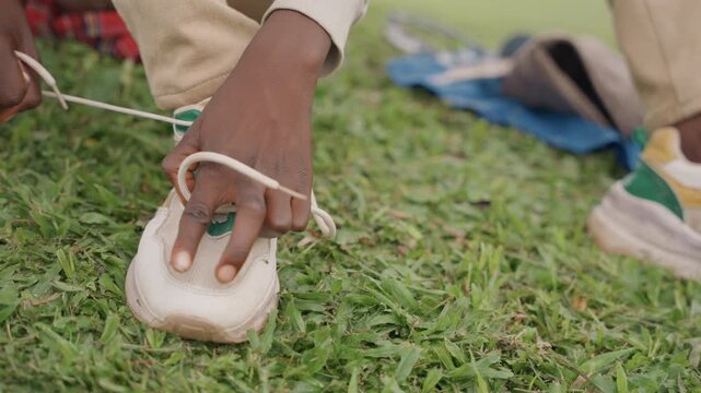 Black father helps son lace sneakers on lawn, closeup hands threading lace through eyelets next to plaid blanket, nurturing lesson in independence and skill, quiet outdoor teaching moment