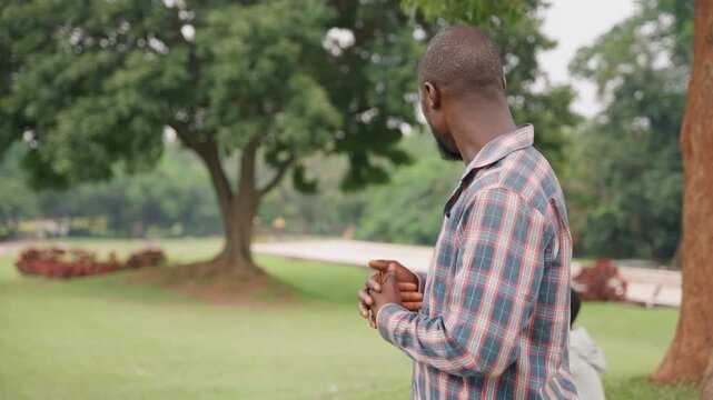 Medium shot black father and son standing under oak in open park, father turning to glance toward son seated on grass, protective posture in patterned shirt, reflective mood and parental