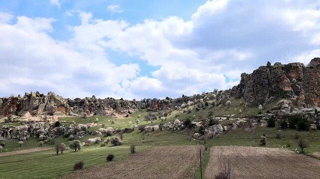 A wide-angle view of Yazılıkaya (Midas City) near Han, Eskişehir, showing the massive Phrygian rock plateau rising above village houses, with spring wheat fields and flowering trees in the foreground,