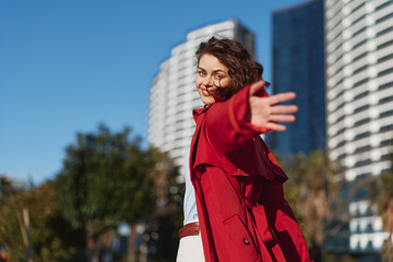 Young woman in a red coat extends her arm toward the camera with a playful smile, standing in a bright urban setting with modern buildings, trees, and blue sky behind. © SHOTPRIME STUDIO