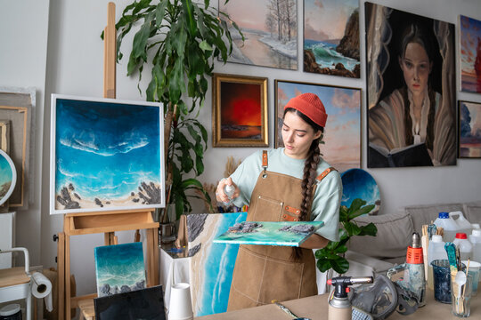 Concentrated craftswoman works on epoxy painting, spraying special cleaning liquid from spray bottle on canvas with resin art to wipe out dust from artwork, focused artist in creative working process.
