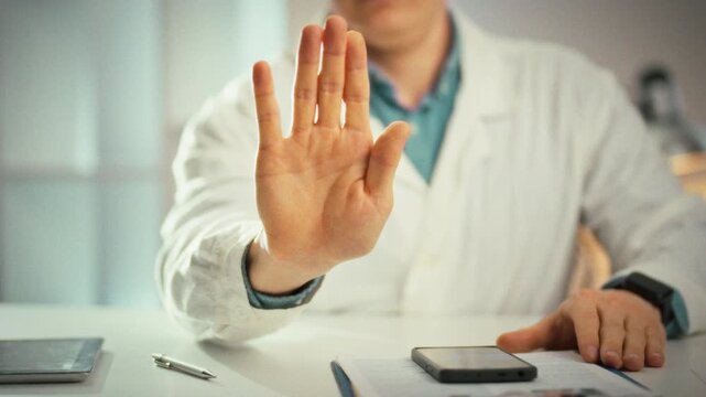 In medical office, a doctor in a crisp white coat raises his hand in a clear stop gesture, emphasizing a medicine-related warning. The close-up highlights the hand, underscoring caution and authority