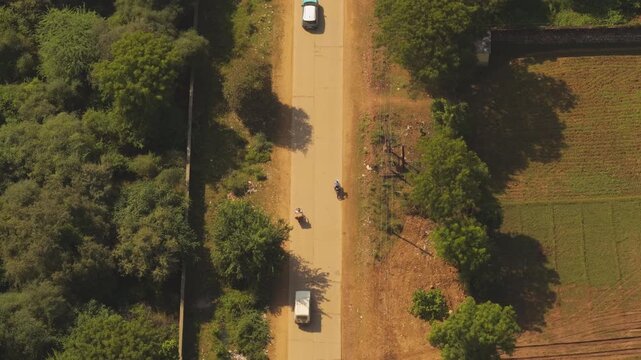 A cinematic highangle shot showing cars and motorbikes traveling along a narrow paved road through the lush green countryside of India during golden hour.