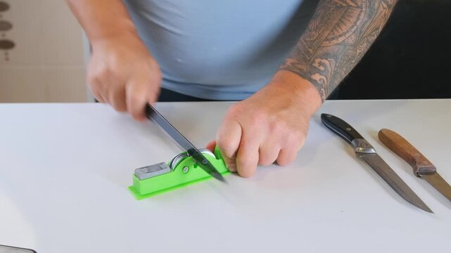 Man sharpening kitchen knife using manual sharpener on table, maintaining blade edge, close view of hands and tools, home kitchen routine, equipment care and safety concept