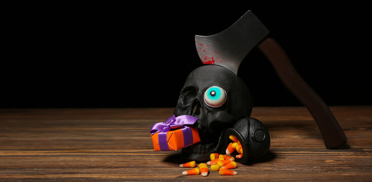Composition with beautiful gift box, tasty candy corns and ax in skull for Halloween on wooden table against dark background, closeup