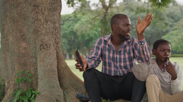Black father and son engaged in animated storytelling under tree, father gesturing with hands, son listening thoughtfully, warm park backdrop, mentorship and wisdom, gentle breeze
