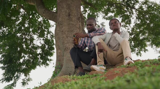 Black father and son looking outward on hillside beneath large tree, father gestures with phone while son watches with hopeful expression, mentoring exchange about dreams and plans, green landscape,