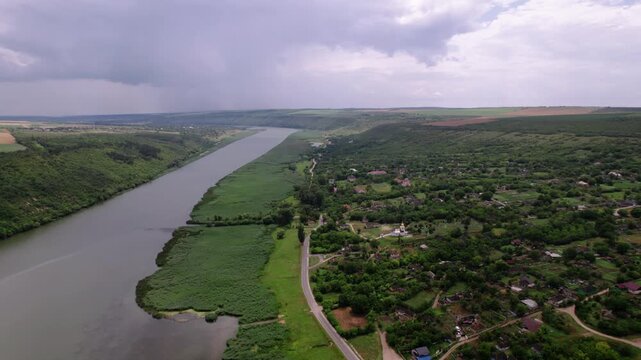 Aerial River Valley Village Under Overcast Sky, Winding River Bordering Green Banks, Scattered Rural Houses And Farm Plots, Unpaved Roads Cutting Through Pastures, Moody Cloudscape