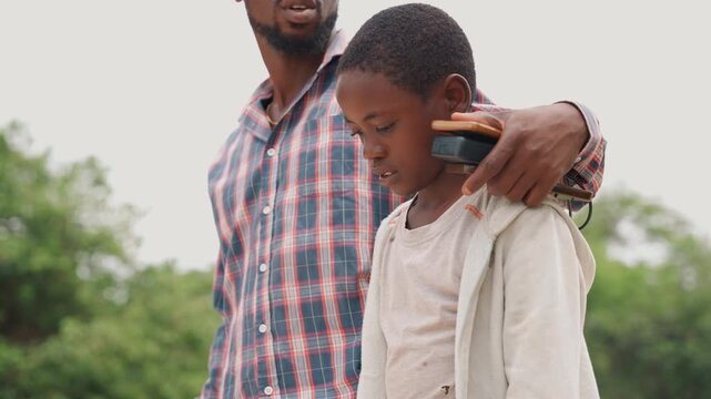 Black father and son close embrace, father with arm around sons shoulder offering comfort and quiet counsel, plaid shirted father guiding son through greenery, tender exchange of reassurance