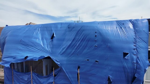 Torn blue tarp flapping in the wind against a building facade. A large blue tarp, ripped and torn by wind, billows dramatically over a building under construction or renovation.