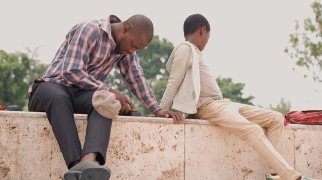 Black father and son on wall in park exhibiting quiet tension as father reaches toward son who leans away muted colors, plaid shirt and jacket, subtle emotion and reflection, generational conversation