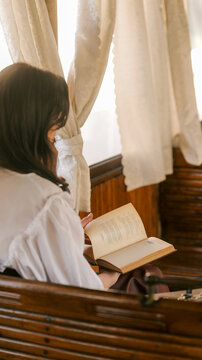 Woman in vintage clothing reading an old book by a window with curtains