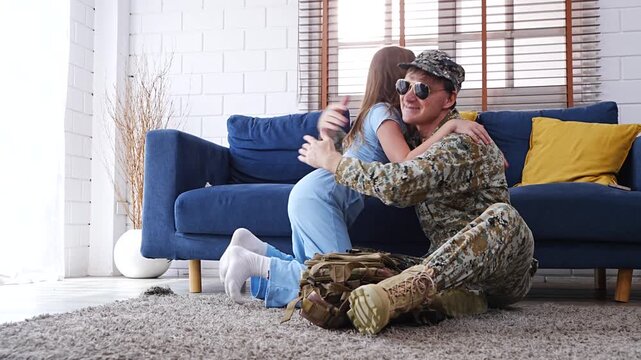 A seven-year-old daughter is helping her father pack his essentials in a backpack as he prepares to wear his military uniform to work before heading to war, in the living room of their house.
