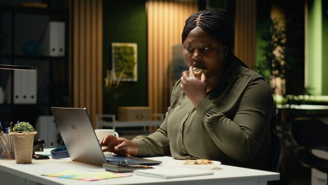African american woman stress eating sweet snack and junk food in the office, showcasing unbalanced work lifestyle situation. Doing overtime and snacking on unhealthy food late at night. Camera B.