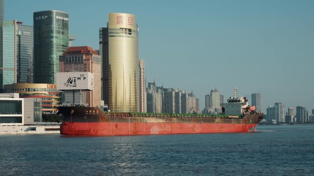 Red cargo ship passing city skyline, golden cylindrical tower and highrise backdrop, calm sea under morning sunlight, industrial port presence, freighter moving along waterfront, urban commerce