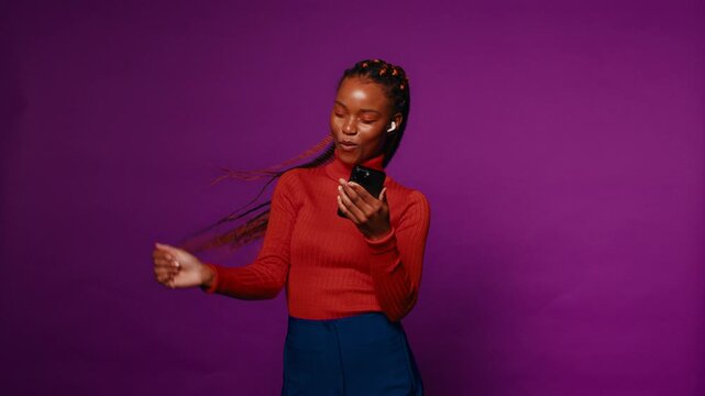 Black woman with colorful braids selects her favorite song on her phone, dancing and singing along to the music while wearing earbuds against a vibrant purple backdrop.
