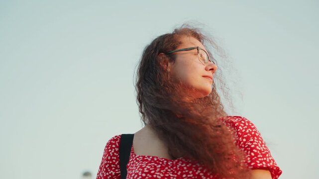 Young woman looking up at skyline during dusk with curly hair swept by wind, wearing glasses and red floral dress, hand brushing hair back in candid motion, tower and city panorama behind her,