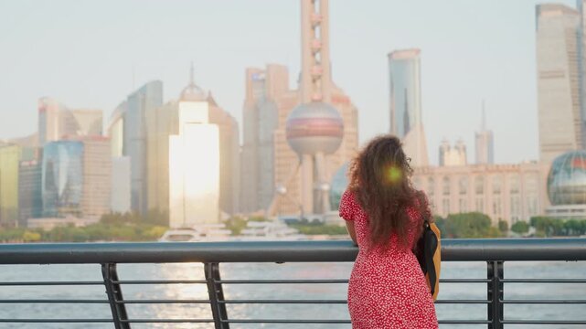 Young woman facing city skyline from waterfront, red dress and backpack, curly hair catching golden sunset, tower reflections on river, urban promenade setting, contemplative travel mood