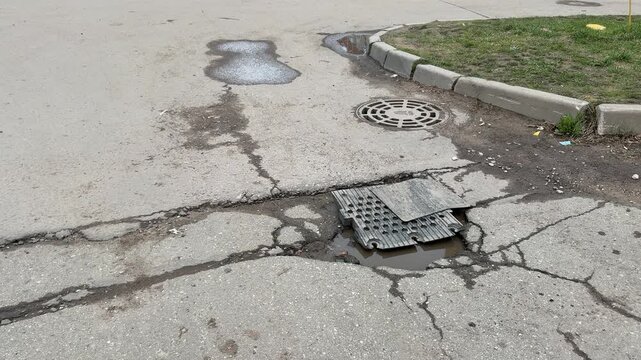 Large pothole on asphalt road covered with improvised metal plate, cracked pavement radiating fractures, shallow water pooling.