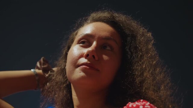 Night rooftop portrait of woman gazing upward with chin raised, rimlit curls, dramatic contrast against dark sky, soft city bokeh, confident aspiration vibe and quiet starlit wonder.