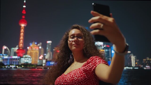 Night skyline selfie with smartphone glow. Curly haired woman in red dress lifts phone for selfie, playful poses and candid smile, glasses catch neon reflections, vlogger and influencer energy,