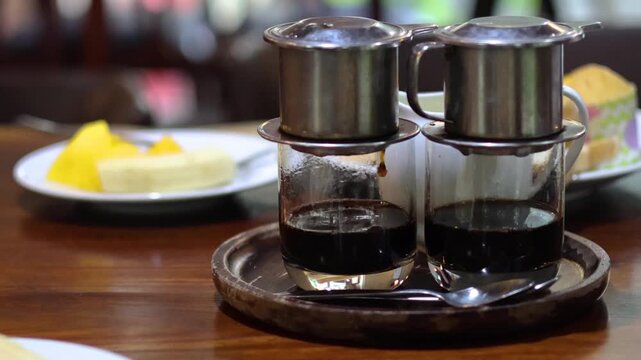 Two cups of traditional Vietnamese phin coffee brewing on a wooden table, with fresh fruit and pastries in background, slow motion shot capturing the process of coffee dripping.