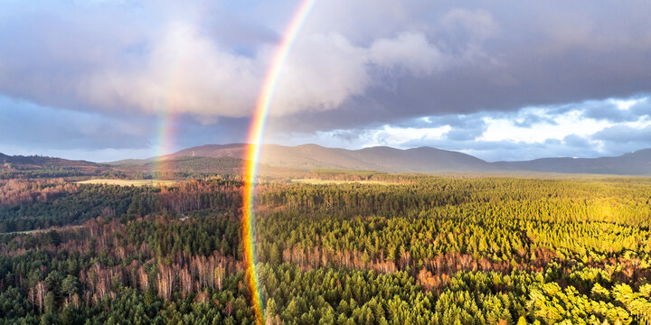 Aerial view of a vibrant double rainbow over the dense evergreen forests and rolling mountains of Cairngorms National Park under a dramatic sky in Scotland, United Kingdom.
