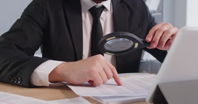 Male auditor wearing formal suit sitting at the desk checking financial documents with magnifying glass. Fraud investigation and tax audit concept. Man accountant searching information. 4k video.