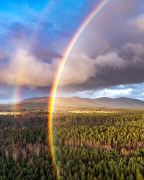 Aerial view of Cairngorms National Park with a vibrant double rainbow over dense evergreen forests and rolling mountains under a dramatic sky in Scotland, United Kingdom.