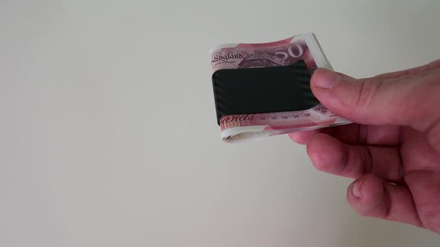 A hand enters from the right in slow motion and brings a sleek black carbon fibre money clip containing several British &pound;50 notes to the centre of frame, holding it steady against a white background.