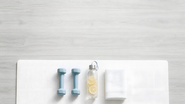 Minimalist home fitness flat lay with white yoga mat, blue dumbbells, and lemon water on grey wood with copy space