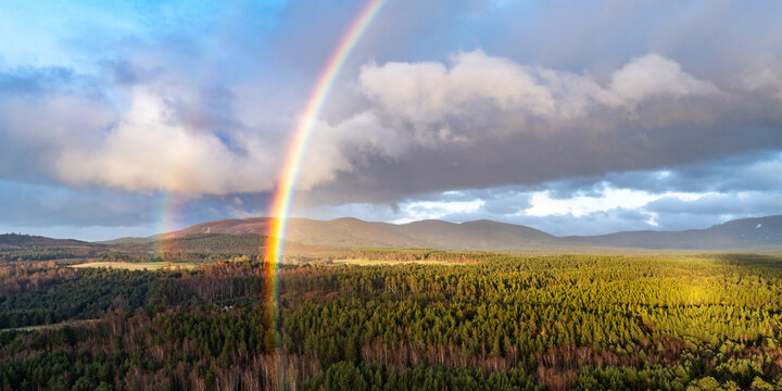 Aerial view of a double rainbow over the dense pine forests and rolling mountains of Cairngorms National Park under a dramatic cloudy sky Scotland, United Kingdom.