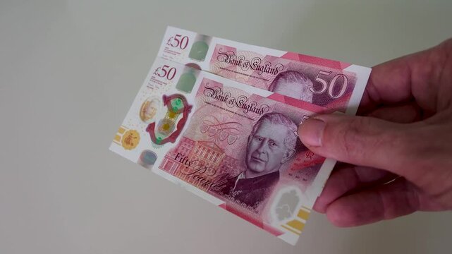 A hand enters frame and brings two British polymer &pound;50 notes to the centre, holding them steady in slow motion against a clean white background, showing King Charles III on the face.