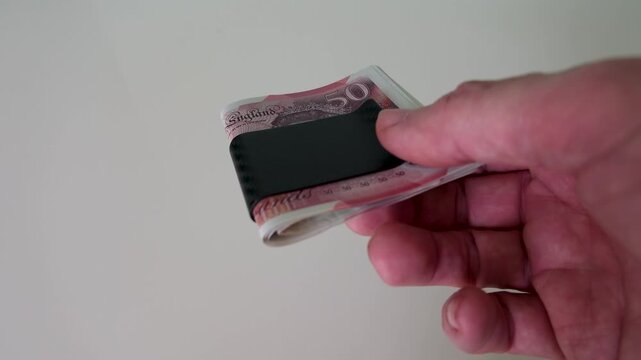 A hand enters from the right holding a sleek black carbon fibre money clip containing several &pound;50 notes, before a second hand enters from the left and takes it smoothly out of frame, slow motion.