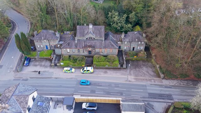 Static drone shot of Windermere police station, traditional Cumbrian slate building, with two liveried police vehicles parked outside as traffic passes on the main road below. Establishing shot.