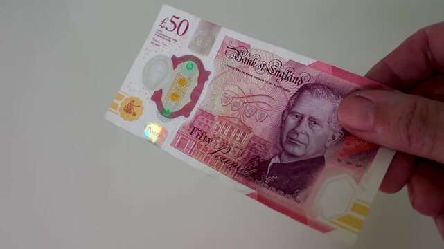 A hand enters from the right holding a British polymer &pound;50 note, holds it centre frame, before a second hand enters and takes the note away to the left, filmed in slow motion against a white backgroun