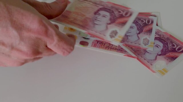 A hand drops a &pound;50 note onto a white surface then methodically counts out a full &pound;1,000 in polymer &pound;50 notes, carelessly building a loosely stacked, dishevelled pile in slow motion.