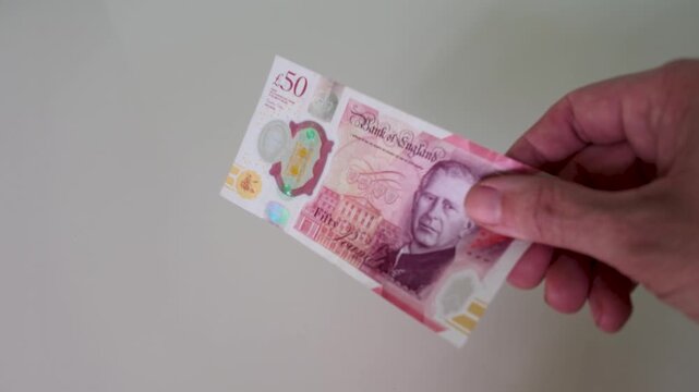 A hand brings a single British polymer &pound;50 note into frame and holds it centrally against a clean white background in ultra slow motion, clearly showing King Charles III on the front.