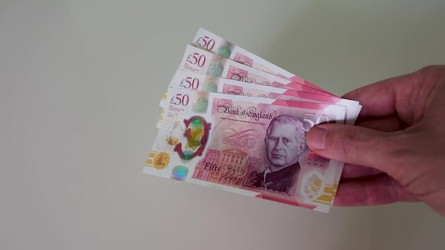 A hand enters from the right holding &pound;200 in British polymer &pound;50 notes with King Charles II, bringing them to centre frame and holding them steady in 120fps slow motion against a white background.
